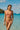 Woman in a pink bikini standing on a beach with clear blue water and a sailboat in the background.