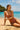 Woman in a pink swimsuit sitting on a sandy beach with clear blue water and sky.