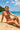 Woman in a pink swimsuit sitting on a beach with clear blue water and sky.