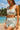 Woman in light blue sports bra and shorts standing on a beach with ocean and sky in the background