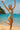 Woman in a green swimsuit jumping on a beach with clear blue water and sky.