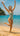 Woman in a green swimsuit jumping on a beach with clear blue water and sky.