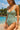 Woman in a green swimsuit standing on a beach with ocean and trees in the background