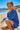 Young girl in a blue patterned outfit sitting on a beach with ocean and mountains in the background.