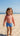 Child wearing a red rash guard and blue swim shorts on a sandy beach.
